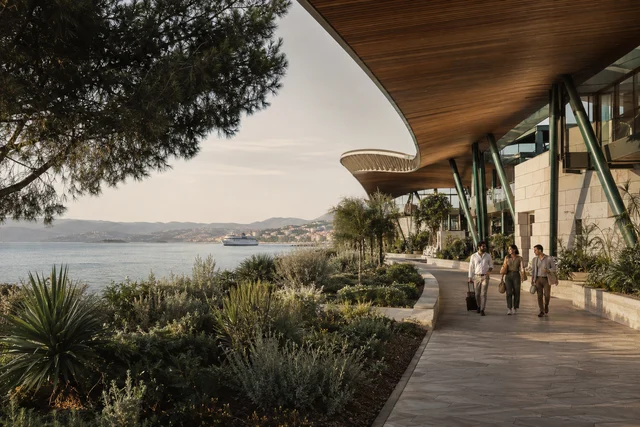 Travellers walking through a planted waterfront terminal with a timber roof overlooking the sea and a coastal city